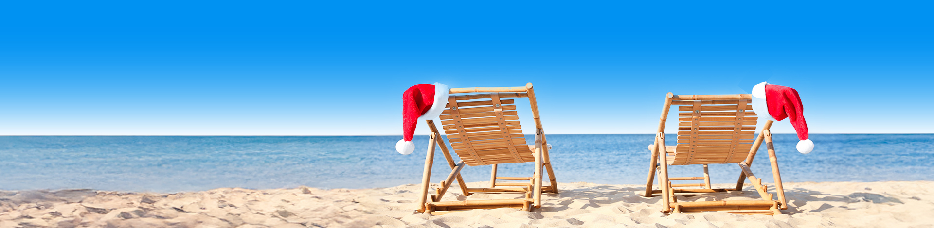 Twee houten strandstoelen met kerstmanmutsen op een zonnig zandstrand, gericht op de kalme blauwe zee onder een heldere lucht.
